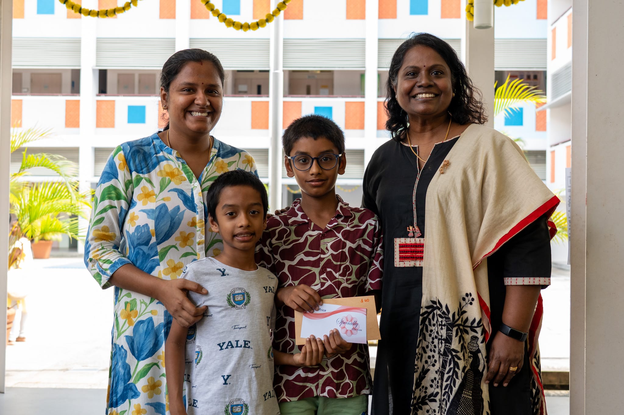 Two women and two boys smile indoors; one boy in glasses holds an envelope, building courtyard behind.