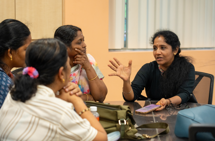 Woman gestures while speaking to three women seated around a table, with bags and a phone on the table.