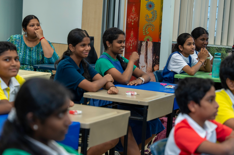Students seated at desks in a classroom, some smiling and leaning forward while listening.