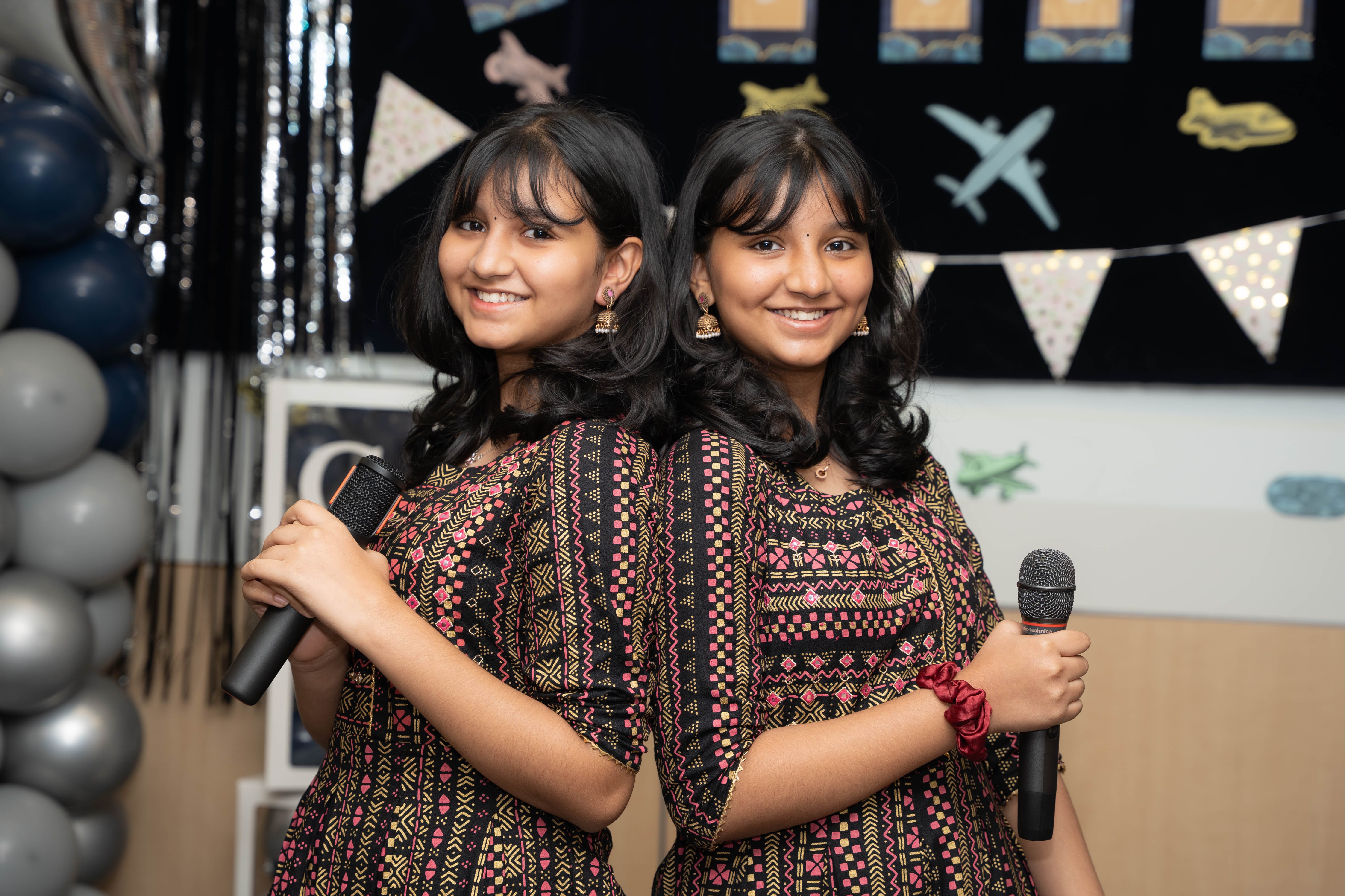 Two girls stand back to back smiling, holding microphones, with balloons and airplane-themed decorations behind.