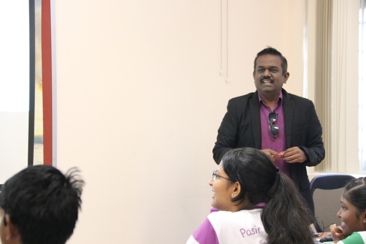 Man in blazer speaks to seated group in a classroom, with a projector screen on the left.
