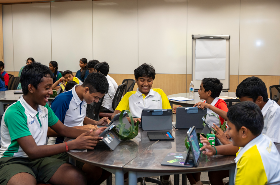 Group of schoolboys in uniforms sit around a table, smiling and using tablets in a classroom.