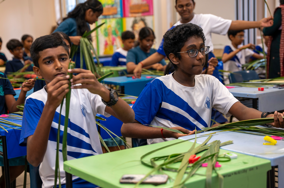 Students in blue-and-white uniforms weave long green leaves at classroom tables during a craft activity.