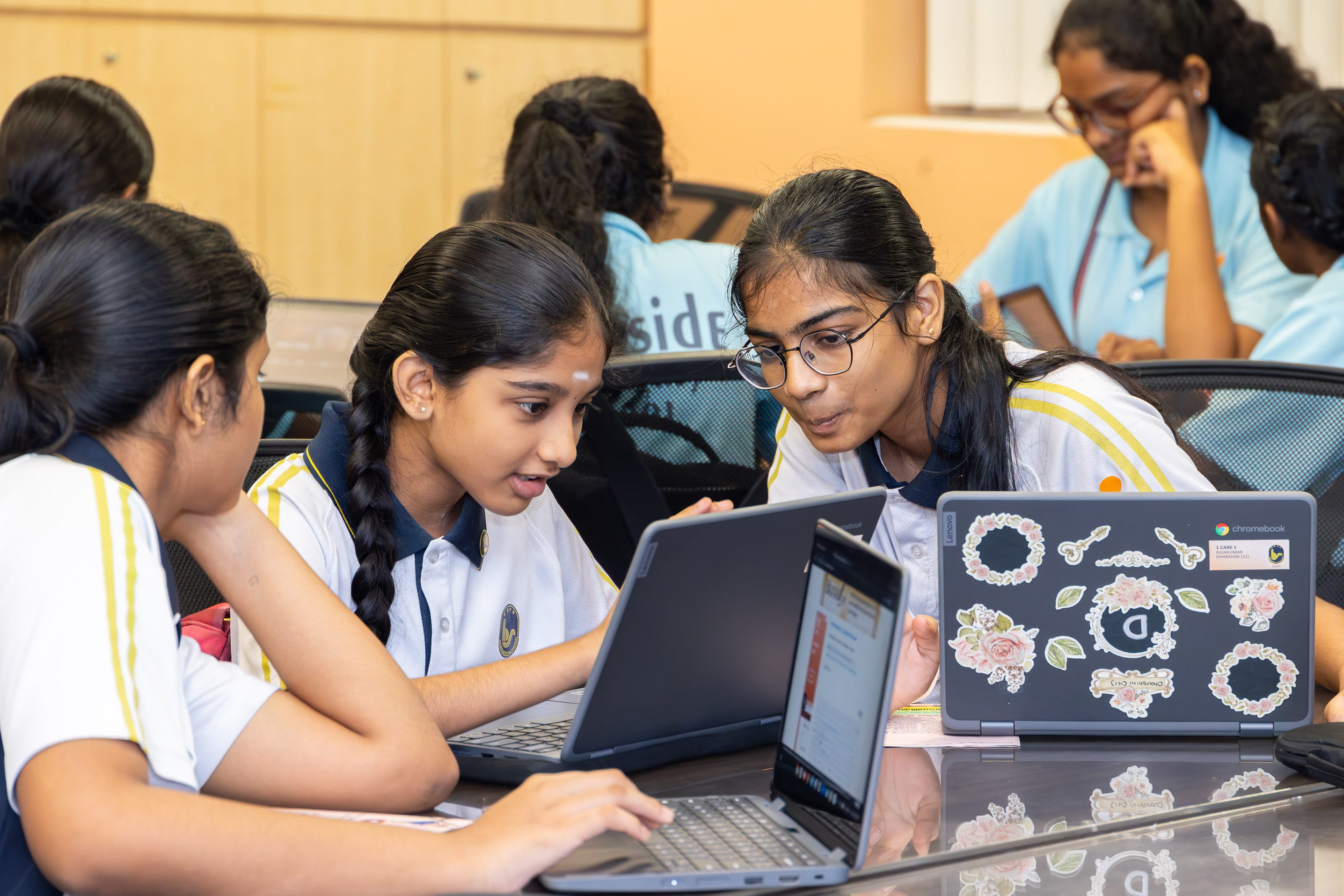 Three students in uniforms huddle over laptops at a table, focused, while others work behind them.