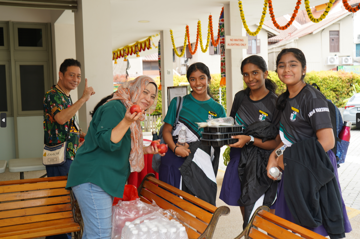 Woman holds apples and smiles with three girls holding food trays; man behind gestures under garlands.