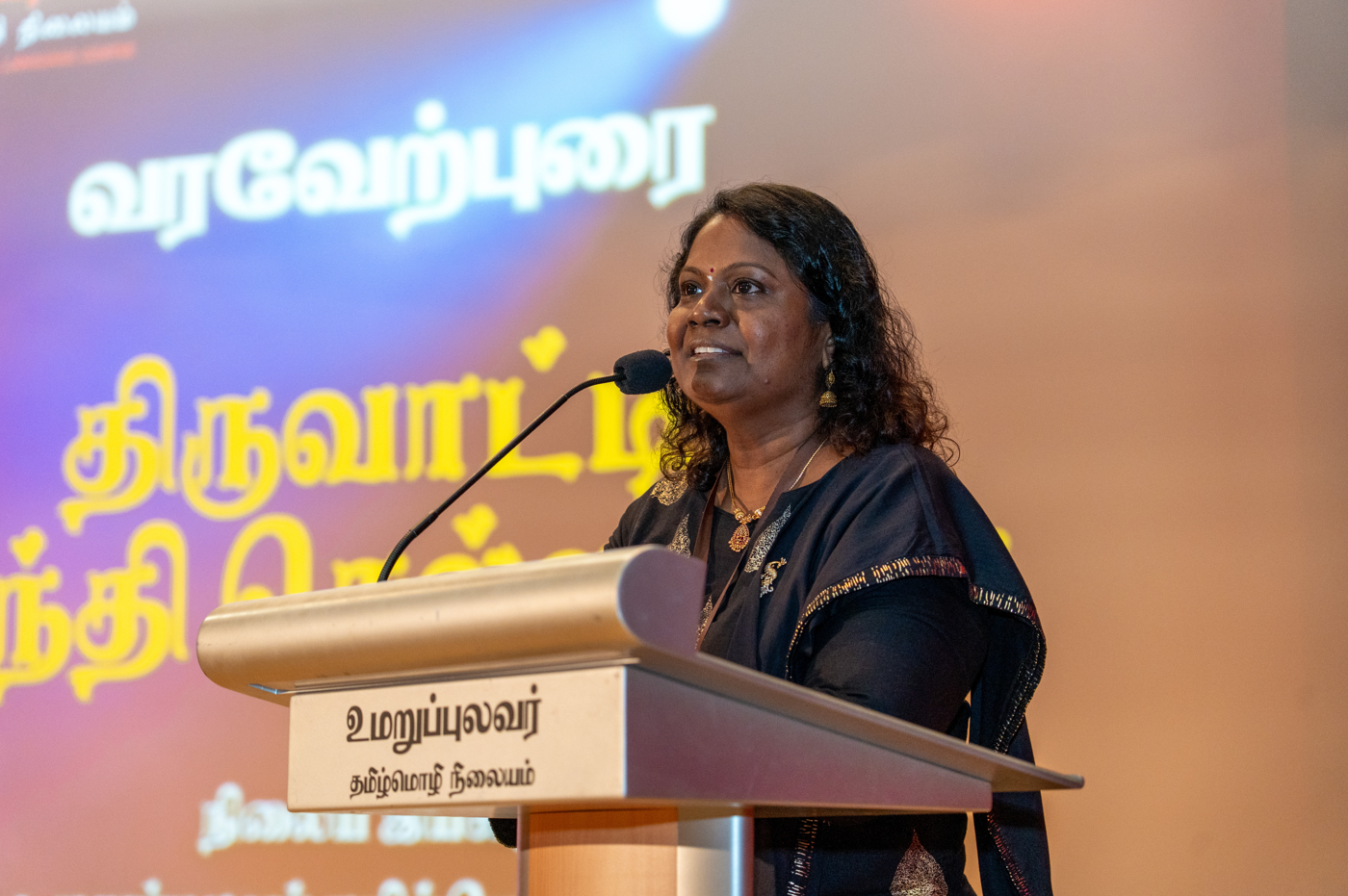 Woman speaks at a podium with a microphone, Tamil text projected on a colorful screen behind her.