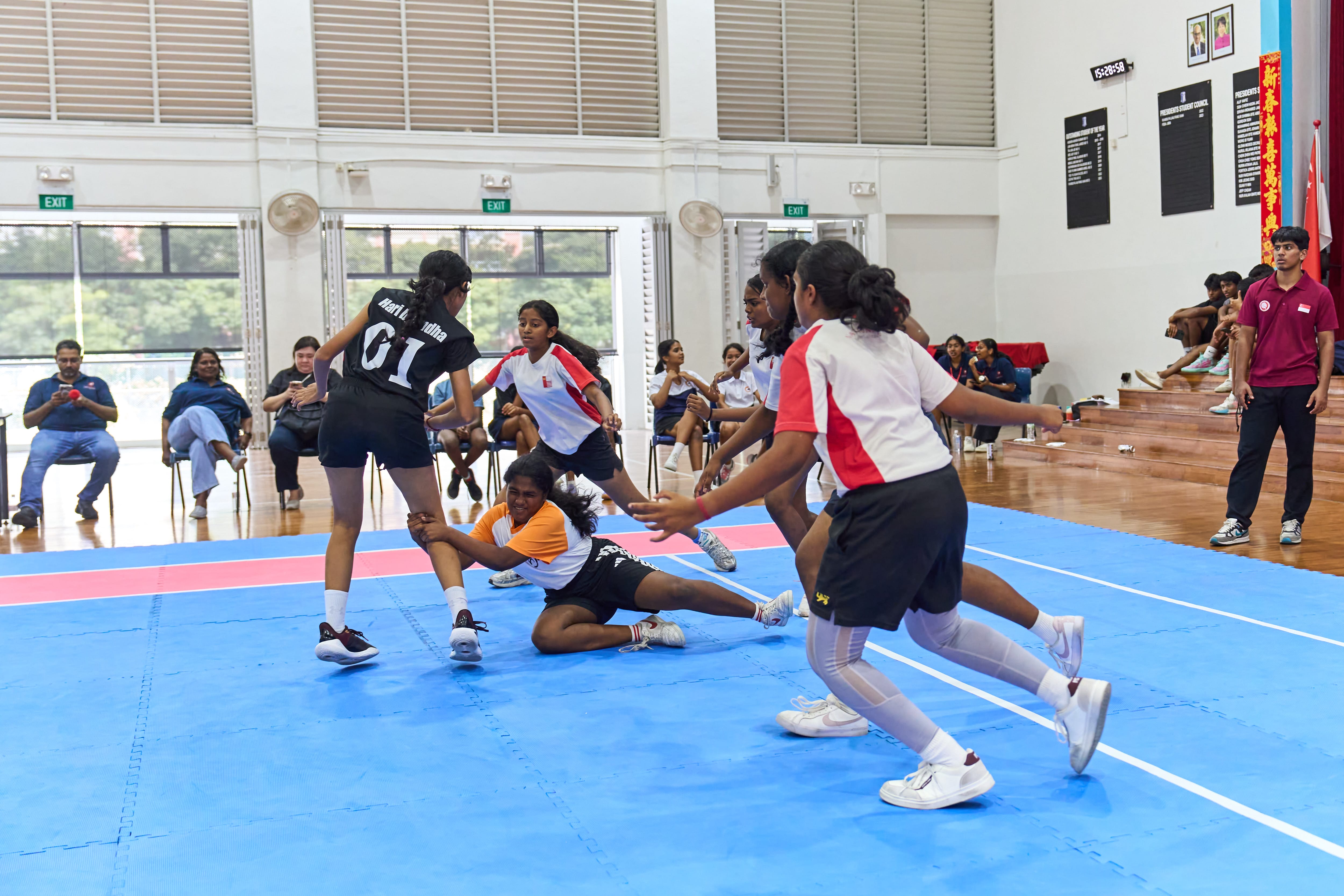 Girls play kabaddi on blue mats indoors as a defender grabs a runner’s leg while others close in.