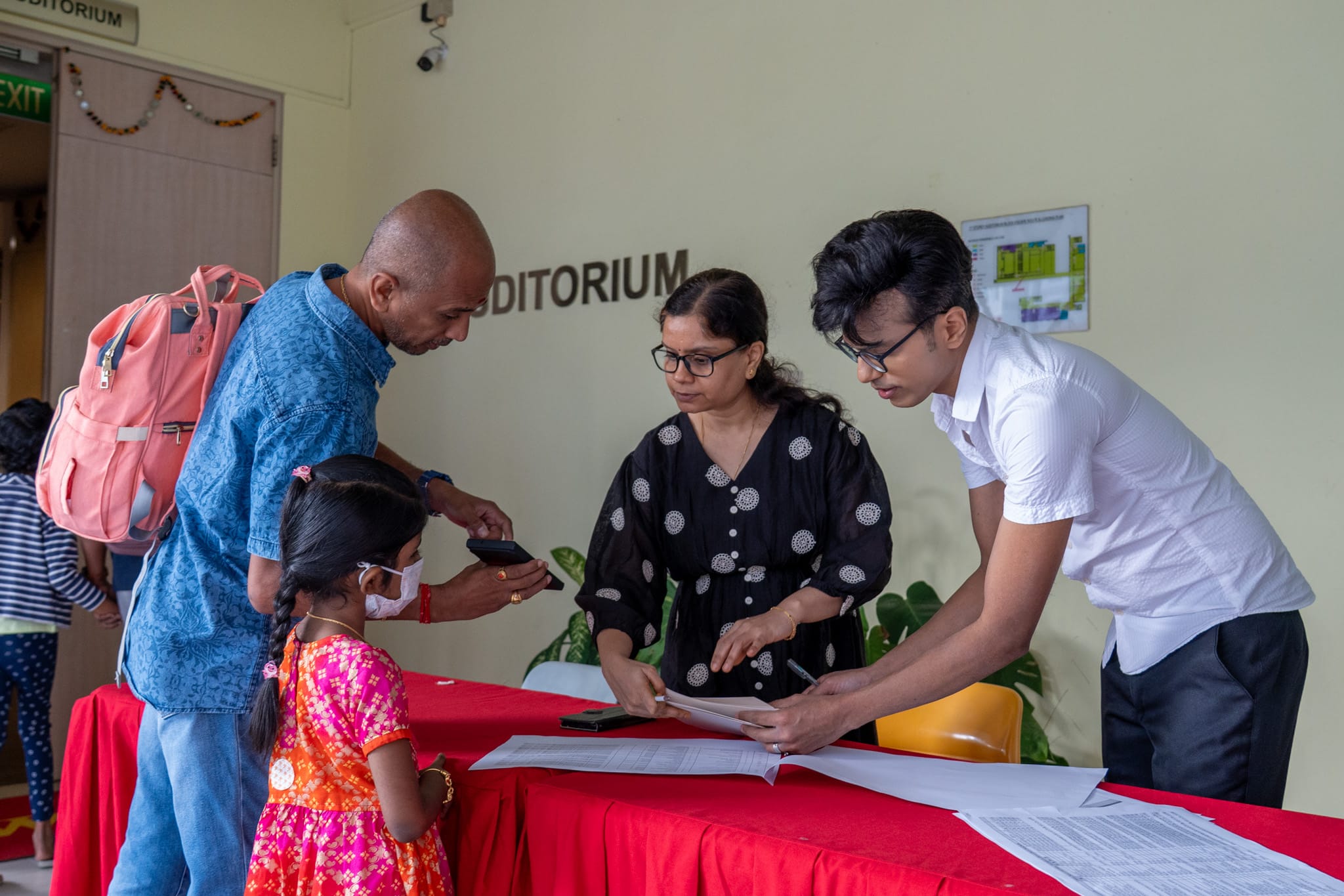 Three adults check papers at a red table while a young girl in a mask watches near an auditorium door.