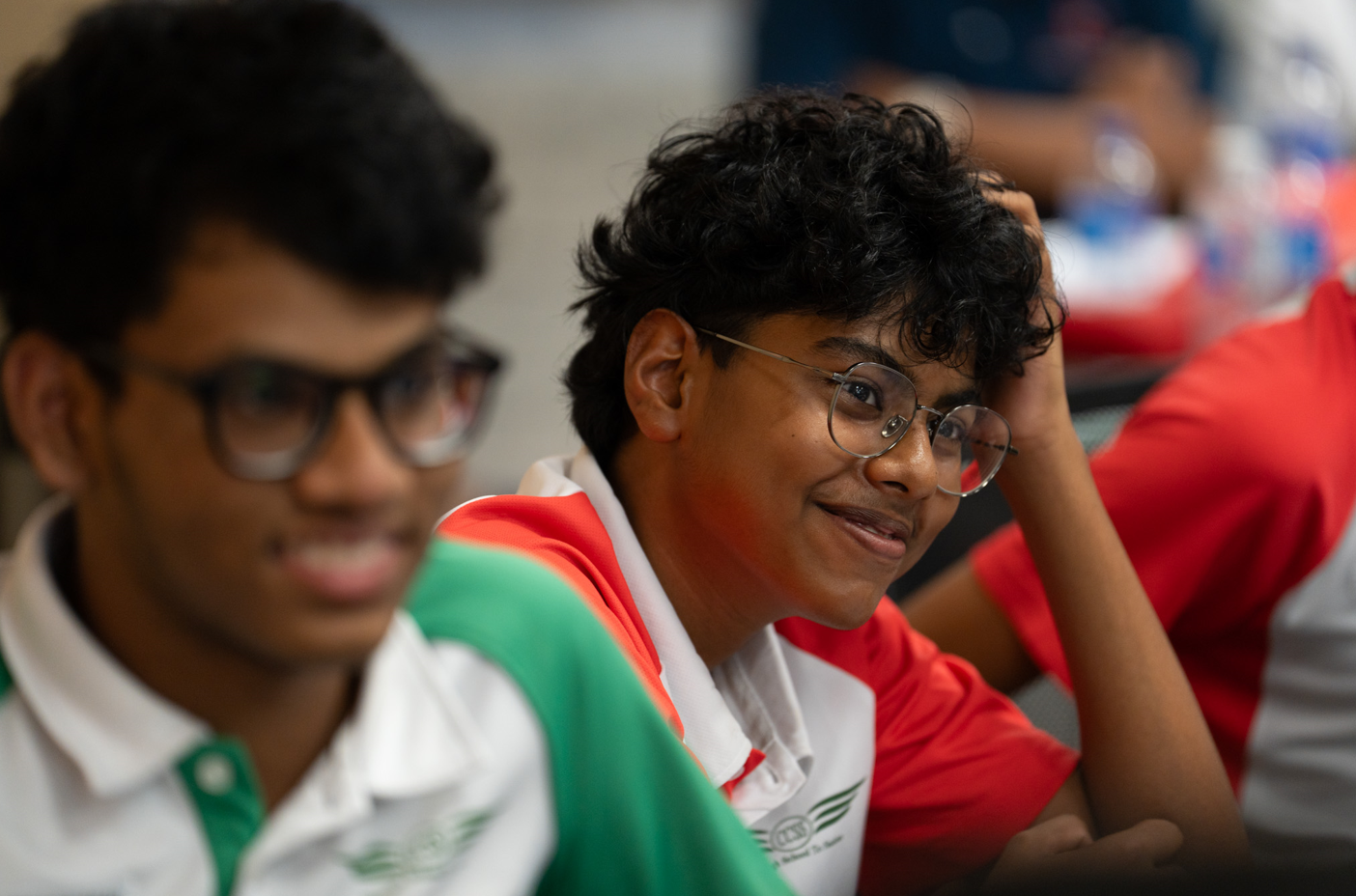 Teen with curly hair and round glasses smiles, leaning on hand, with another student blurred in foreground.