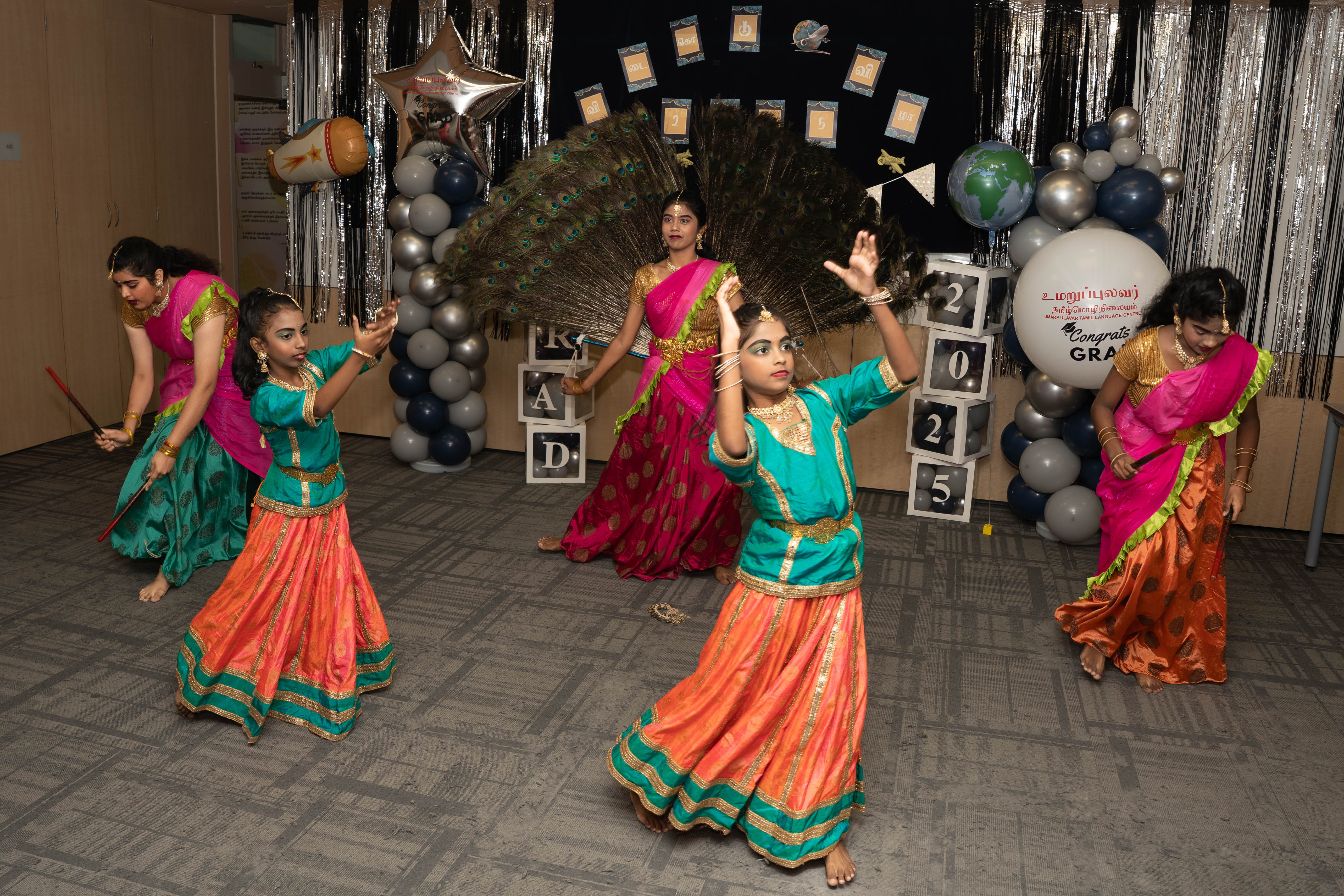 Four girls in bright traditional dresses dance on a decorated stage with balloons and silver streamers.