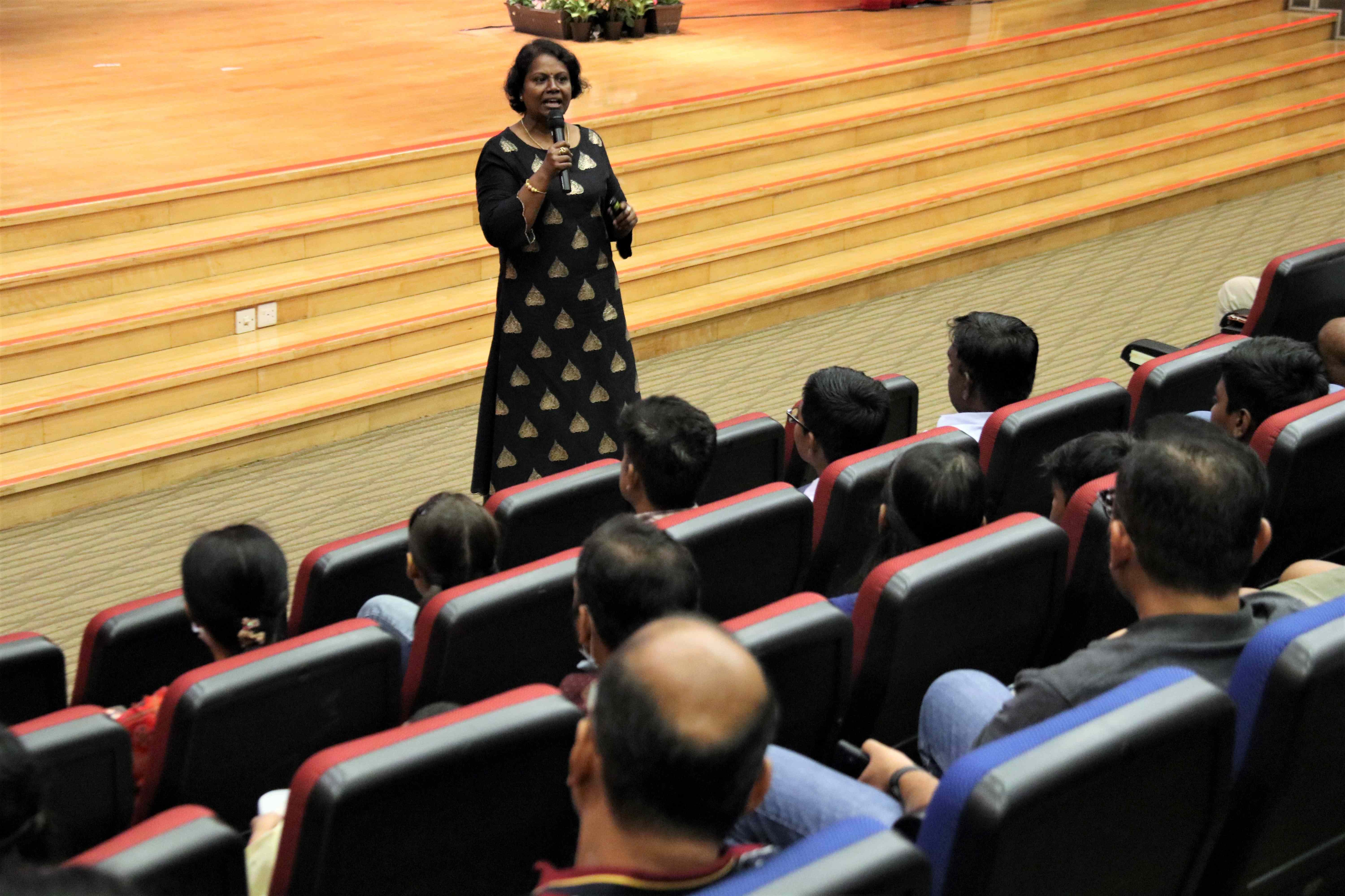 Woman with microphone speaks to seated audience in an auditorium, standing near wide wooden steps.