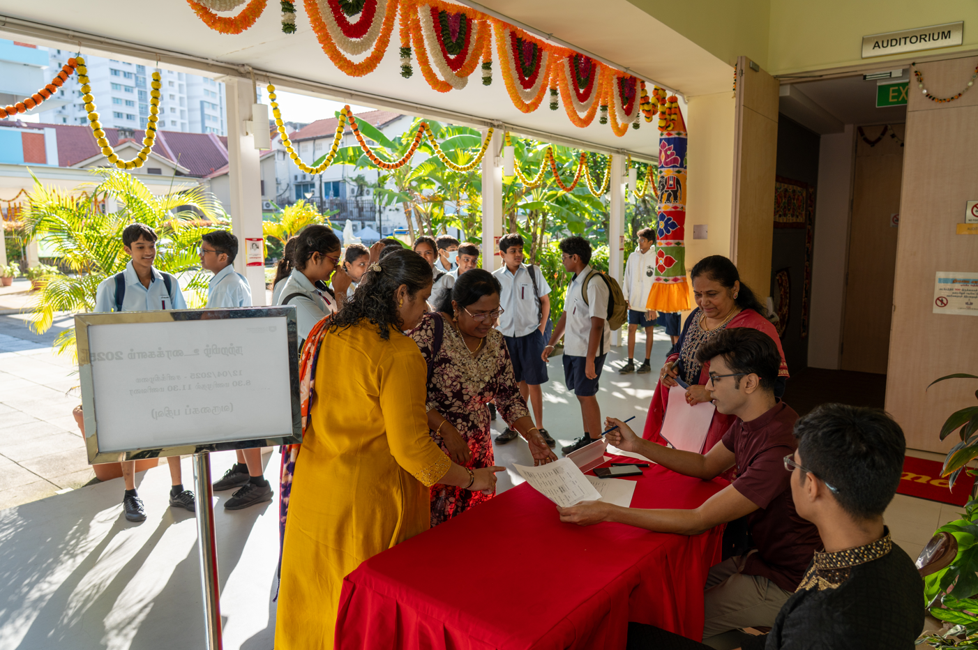 People register at a red table under floral garlands while students in uniforms gather in a corridor.