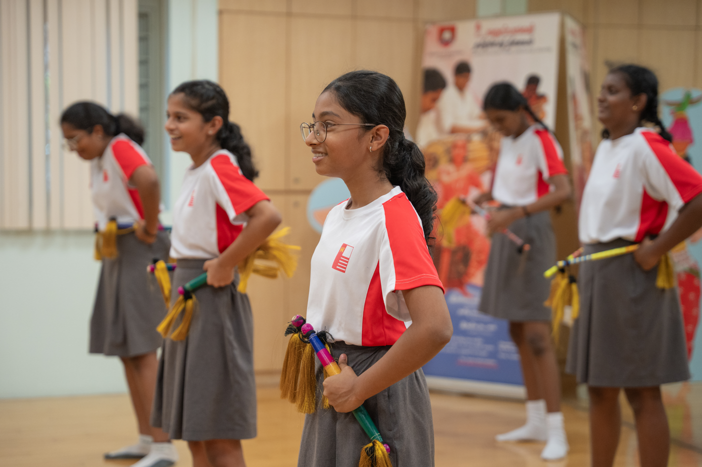Schoolgirls in white-and-red shirts and gray skirts hold colorful sticks, standing in a line indoors.