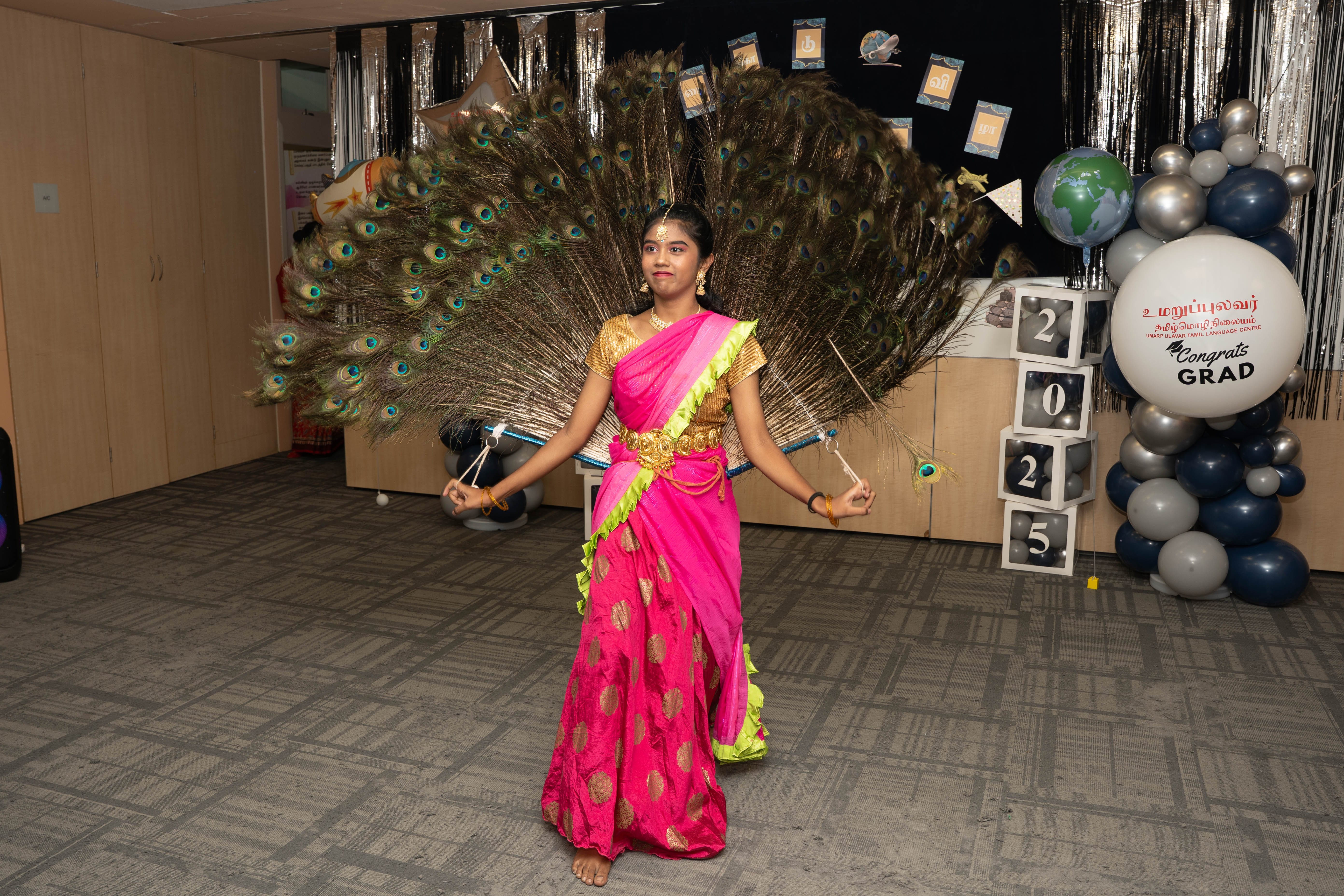 Dancer in bright pink sari holds a huge fan of peacock feathers, standing at a graduation-themed backdrop.