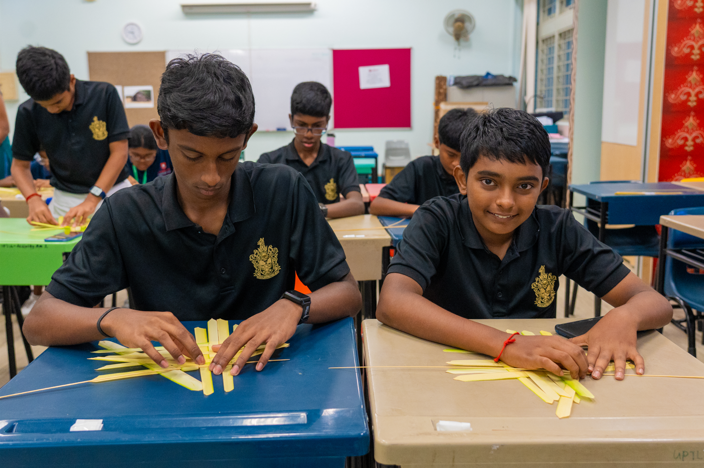 Students in black uniforms weave pale yellow strips at desks in a classroom; one boy smiles at camera.