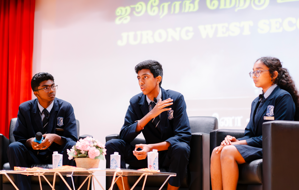 Three students in school uniforms sit on stage chairs; one speaks with hand raised, another holds a microphone.