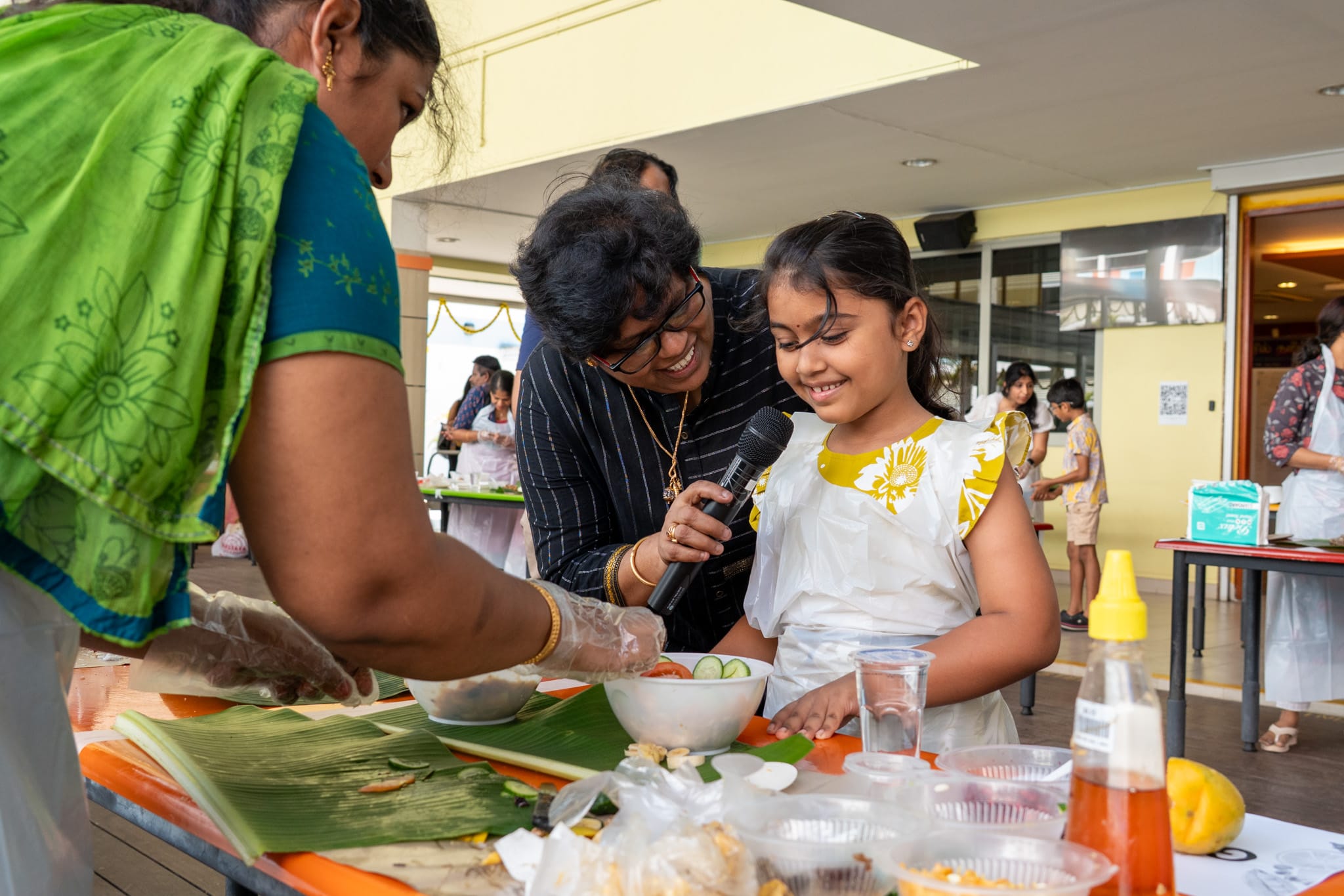 Man holds a microphone to a smiling girl in apron as a woman prepares food at a table with bowls and leaves.
