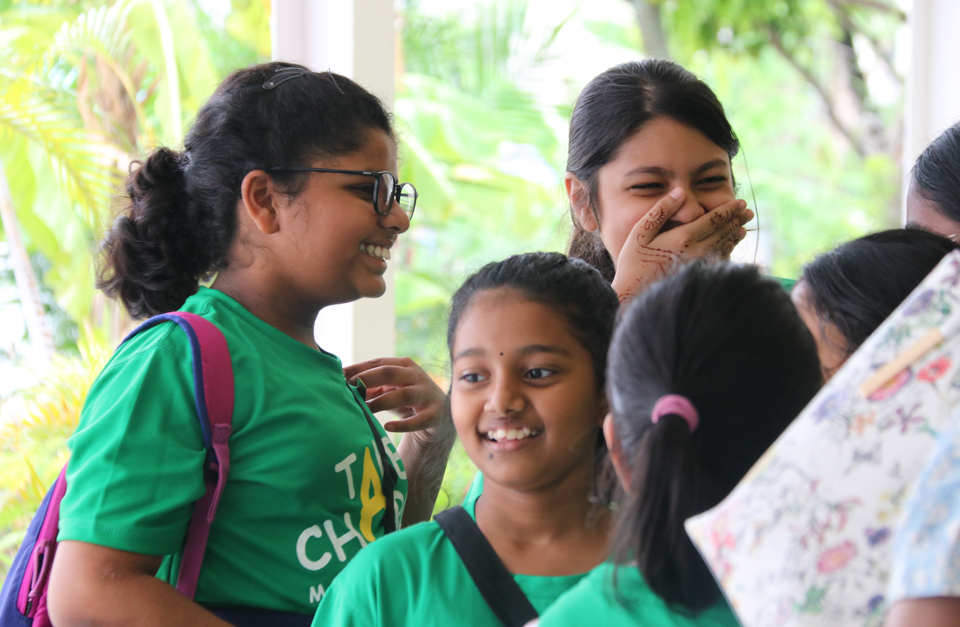 Group of girls in green shirts smiling and laughing, one covering her mouth, outdoors with greenery behind.