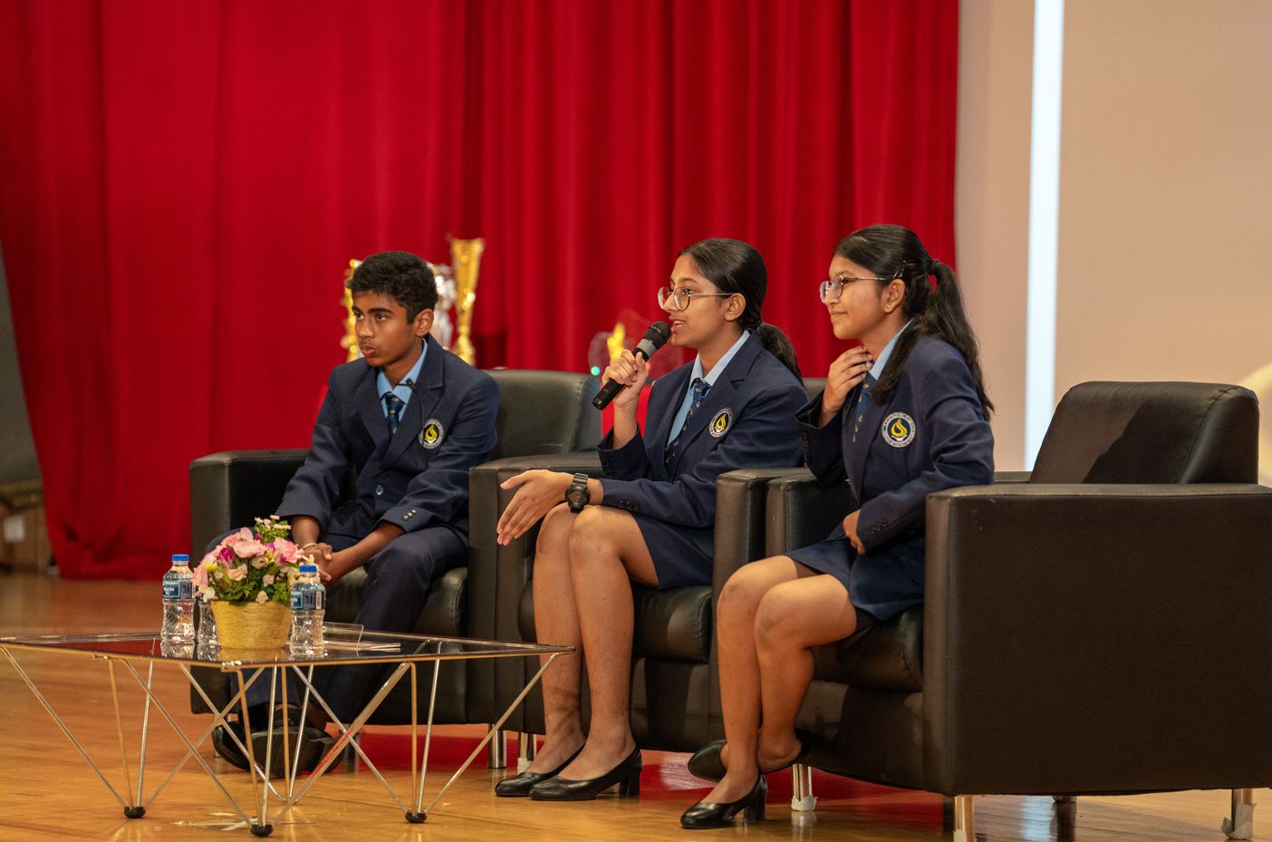 Three students in navy uniforms sit on black armchairs; one speaks into a microphone before red curtains.