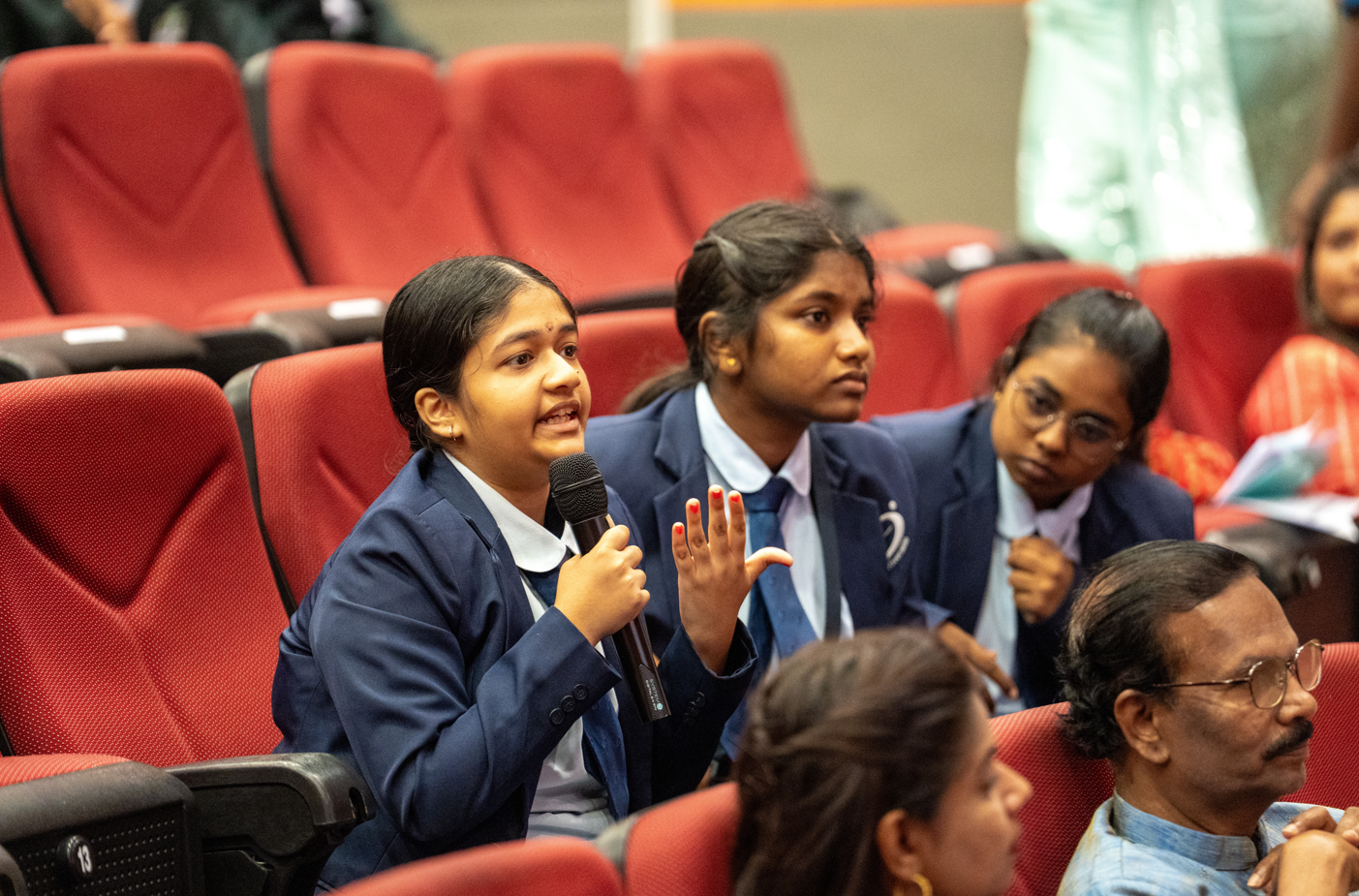 Student in blue blazer speaks into a microphone, hand raised, seated among others in red auditorium seats.