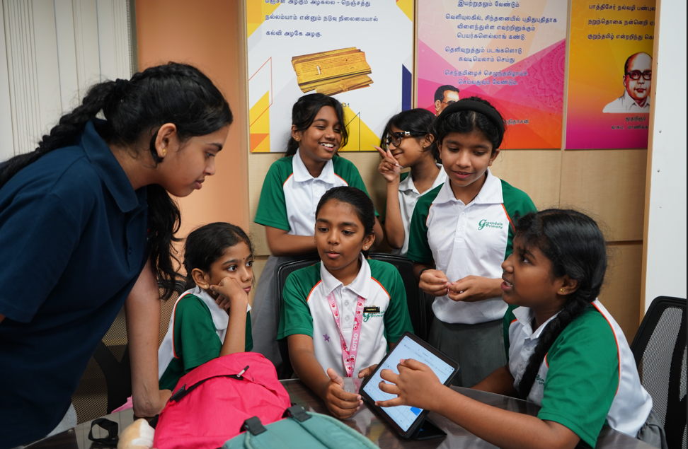 Teacher leans over a table as six schoolgirls in green uniforms talk and look at a tablet.