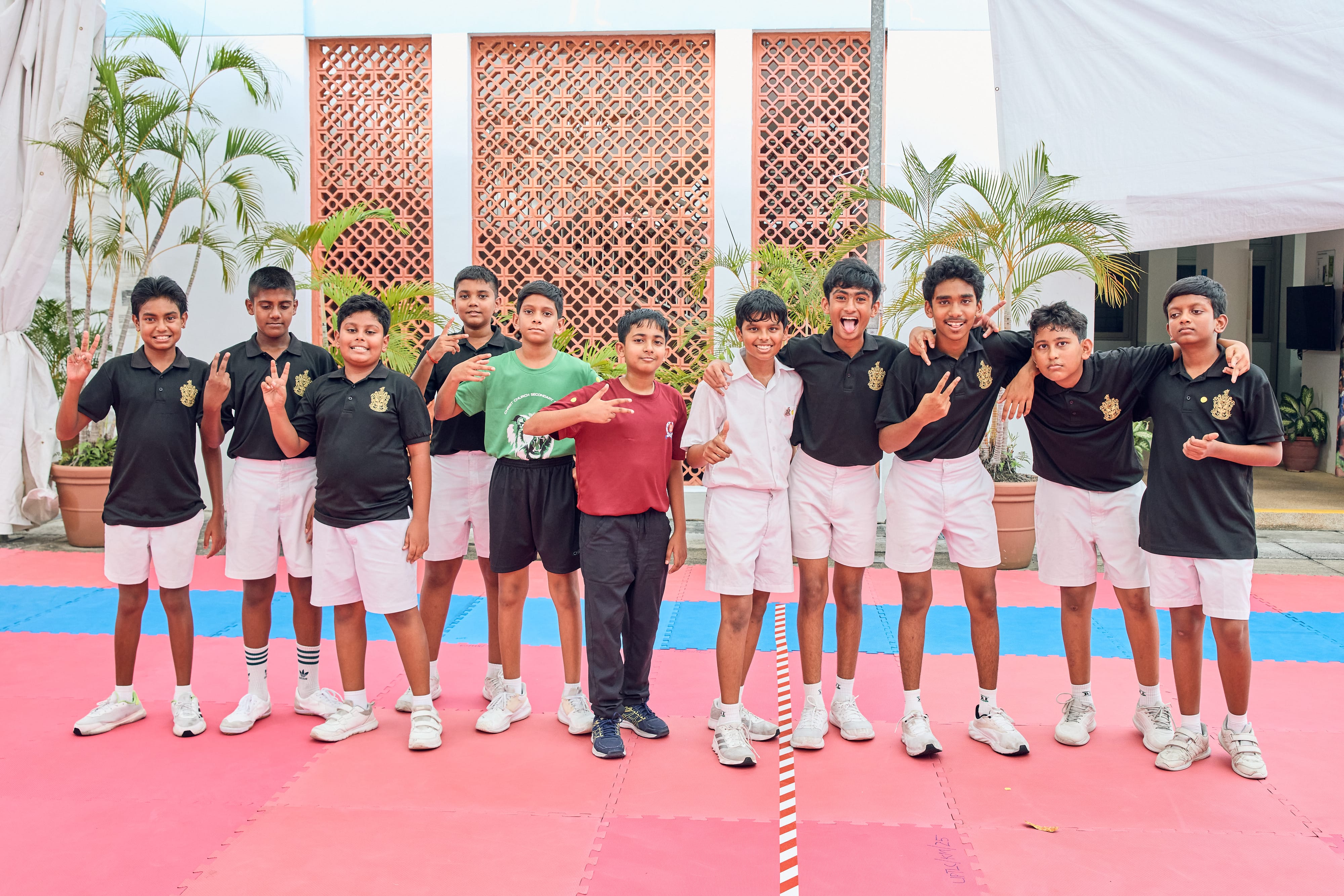 Eleven boys pose on red and blue mats outdoors, some making peace signs, with palm plants behind.