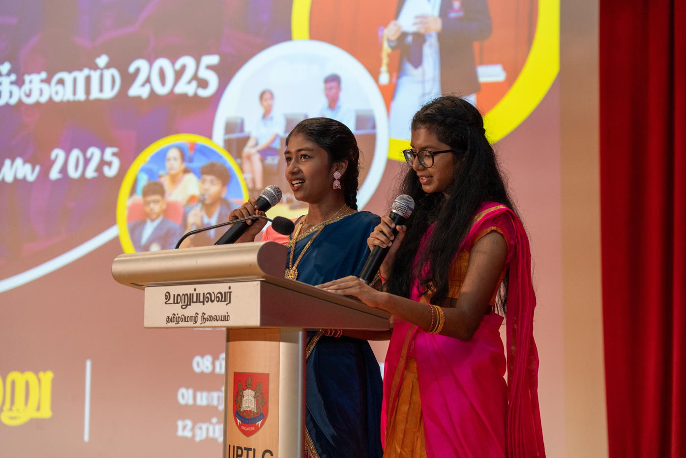 Two girls in colorful saris speak into microphones at a podium, with a 2025 event screen behind.