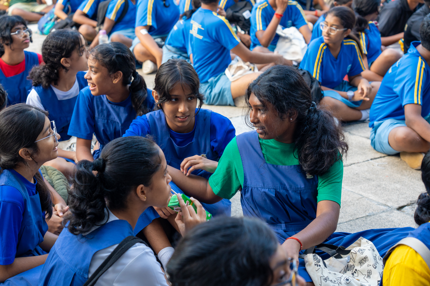 Students in blue uniforms sit on pavement in groups, chatting; one in green shirt talks to a friend.