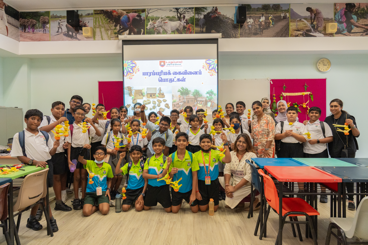 Group of students and teachers posing in a classroom, holding yellow flower garlands before a projector screen.