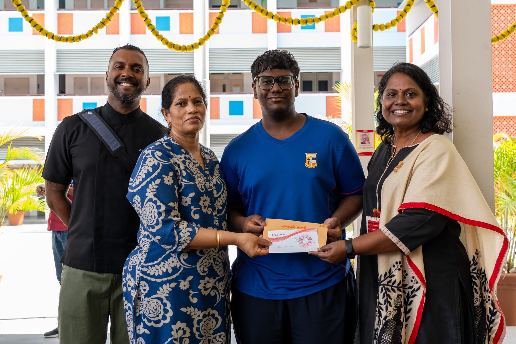 Four people pose indoors as a teen holds an envelope, with marigold garlands hanging above.