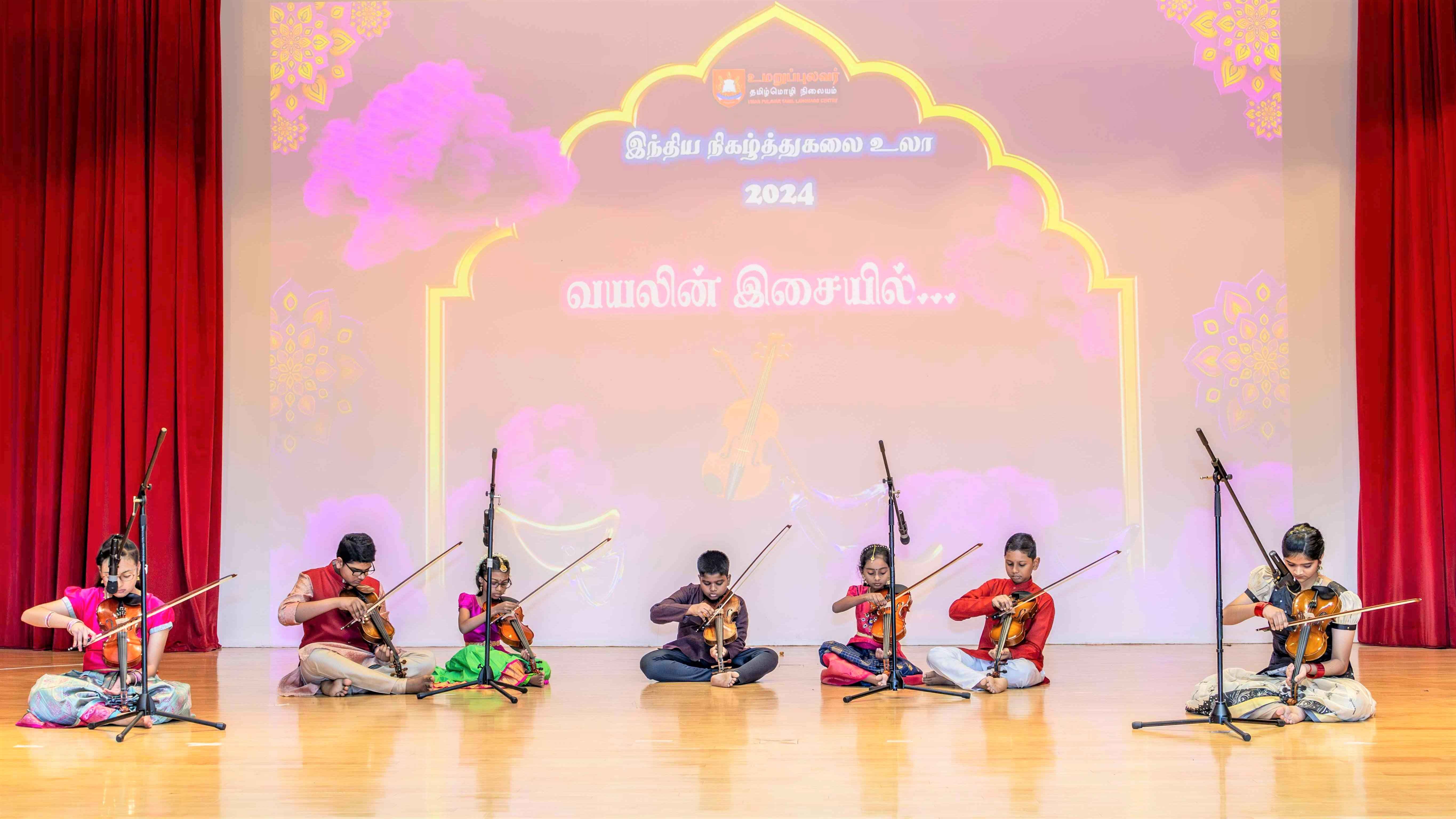 Seven children sit onstage playing violins, with microphones and Tamil text projected behind them.