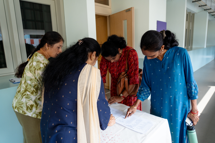 Four women lean over a table, signing or reviewing papers in a bright hallway.