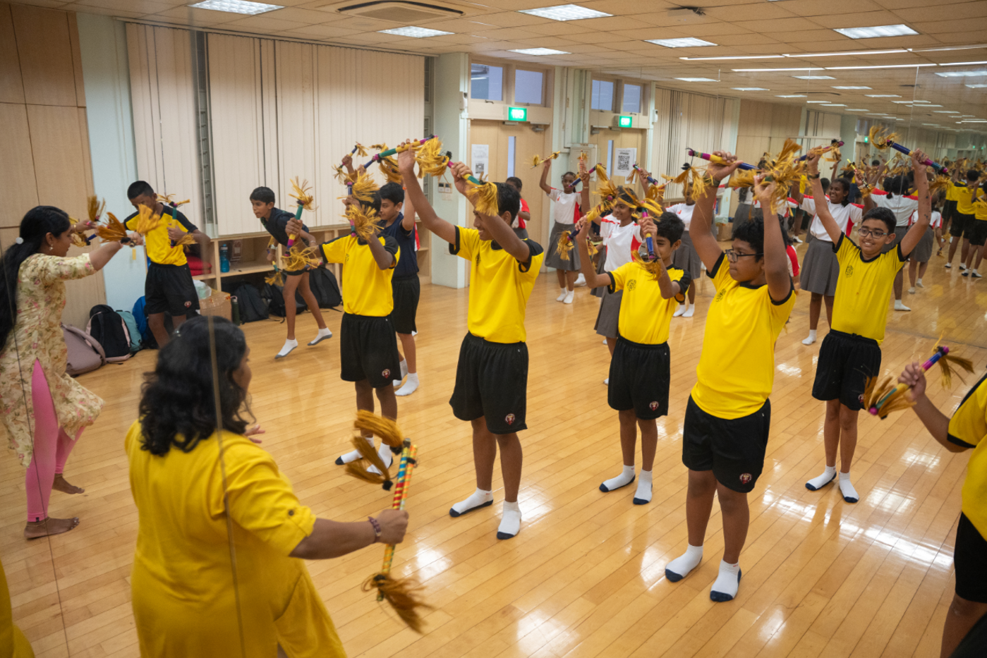 Students in yellow uniforms practice a stick-and-tassel dance in a mirrored studio with an instructor.