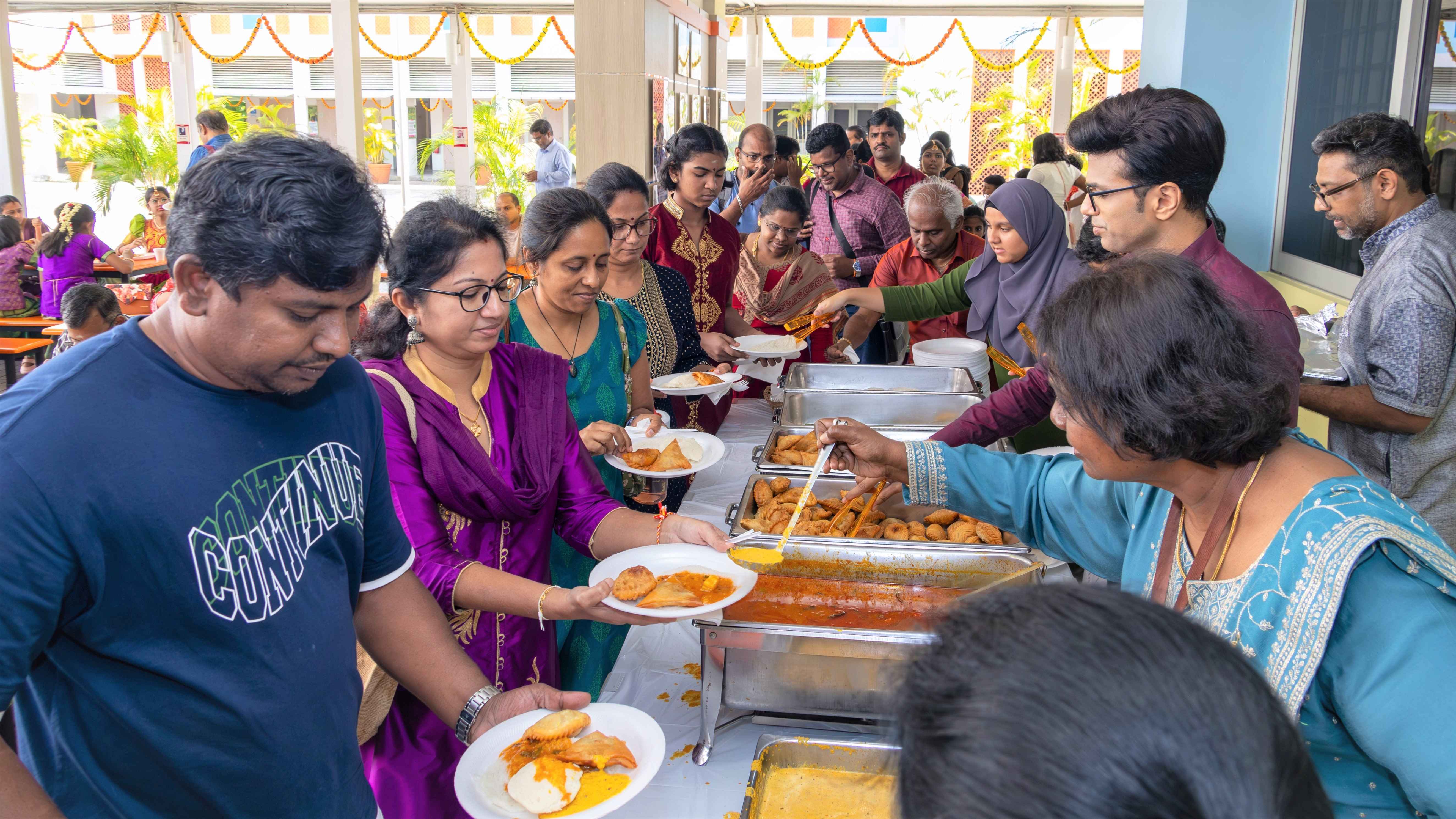 People in festive clothes line up at a buffet while servers dish curry and fried snacks onto plates.