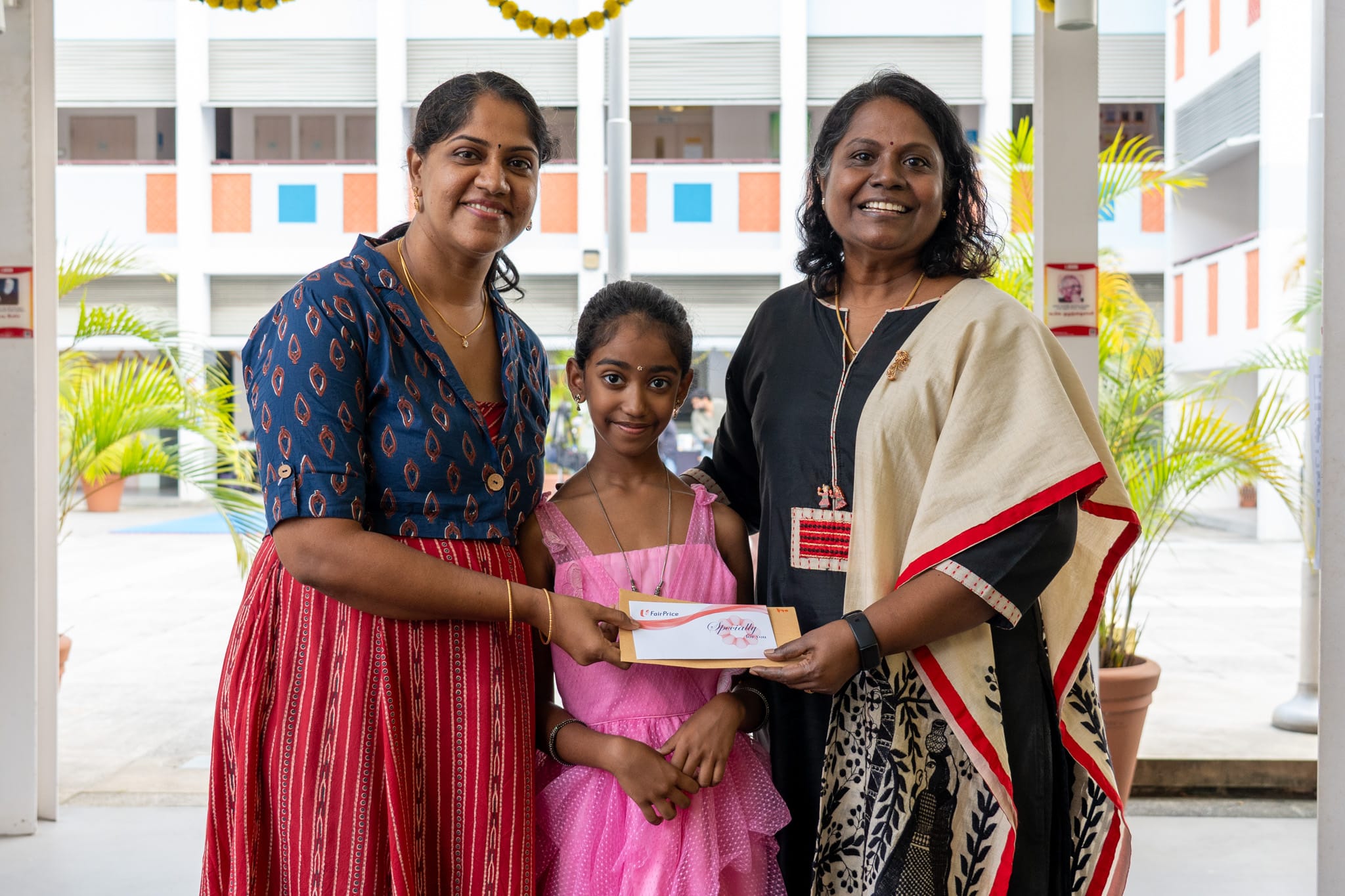 Two women and a girl smile while holding a certificate in a courtyard with potted plants behind them.