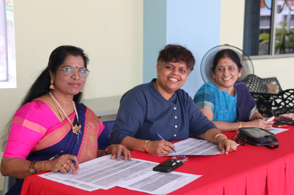 Three women sit at a red-covered table, smiling and filling out forms with pens.