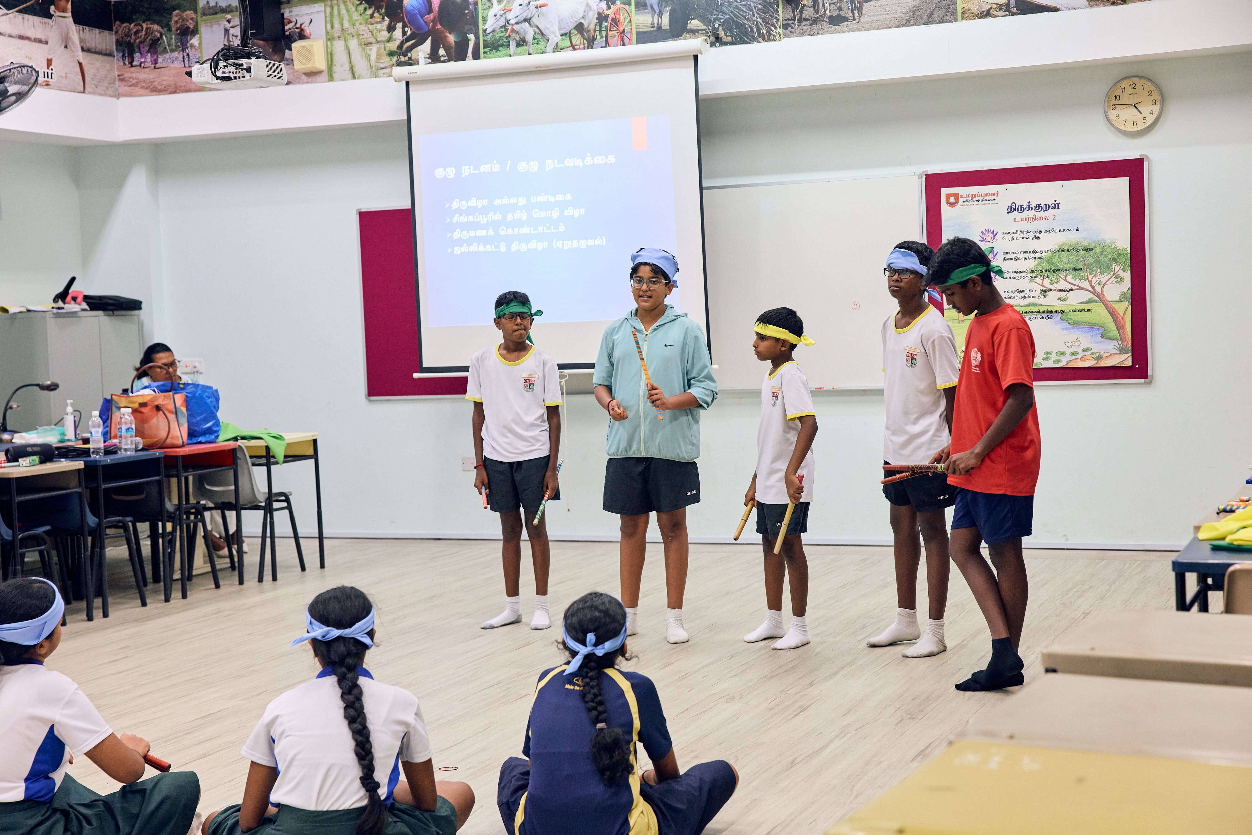 Five students with blindfolds stand in a classroom holding sticks, presenting to seated classmates.