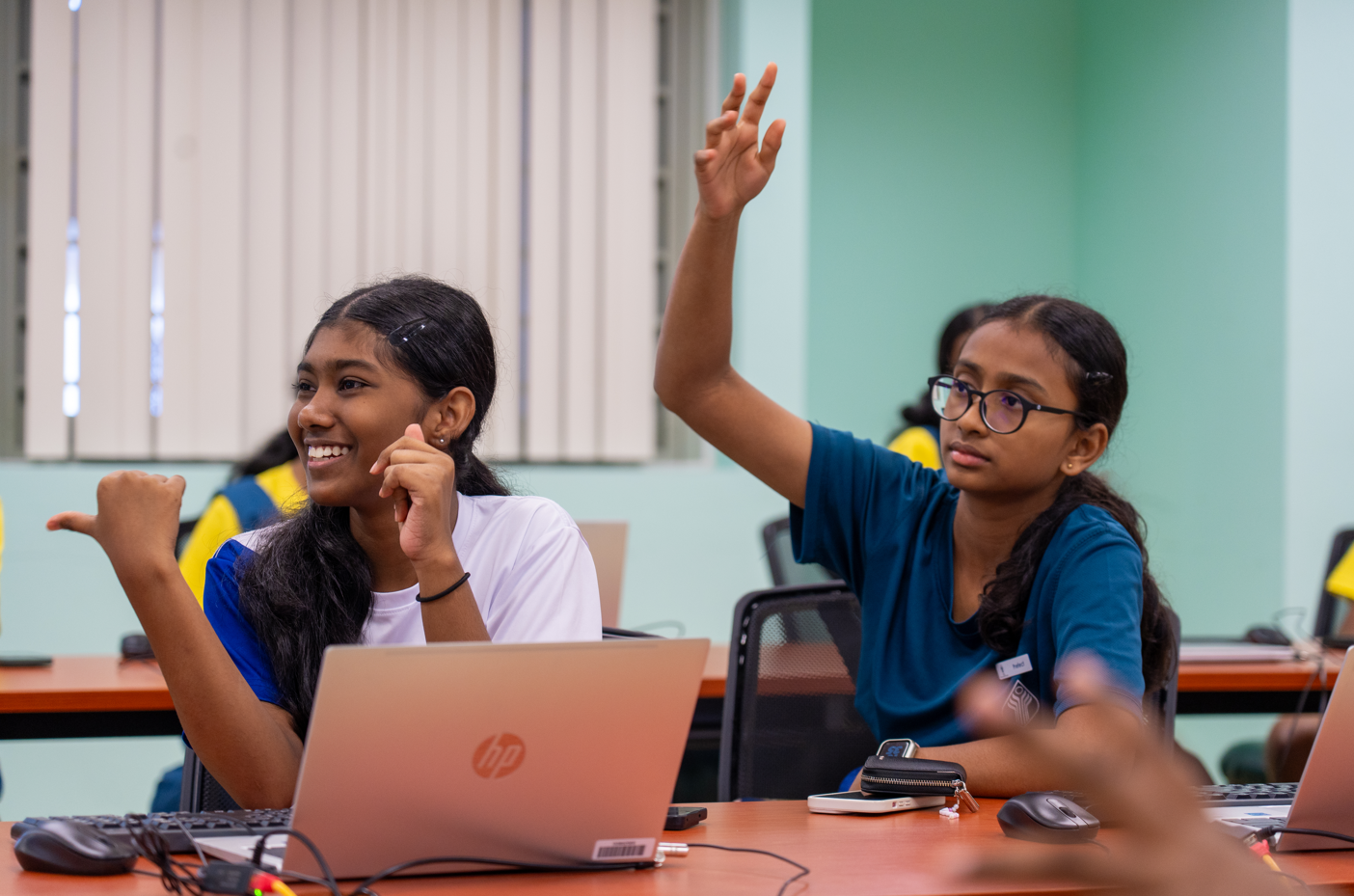 Two students at desks with laptops; one smiles while the other, wearing glasses, raises her hand.