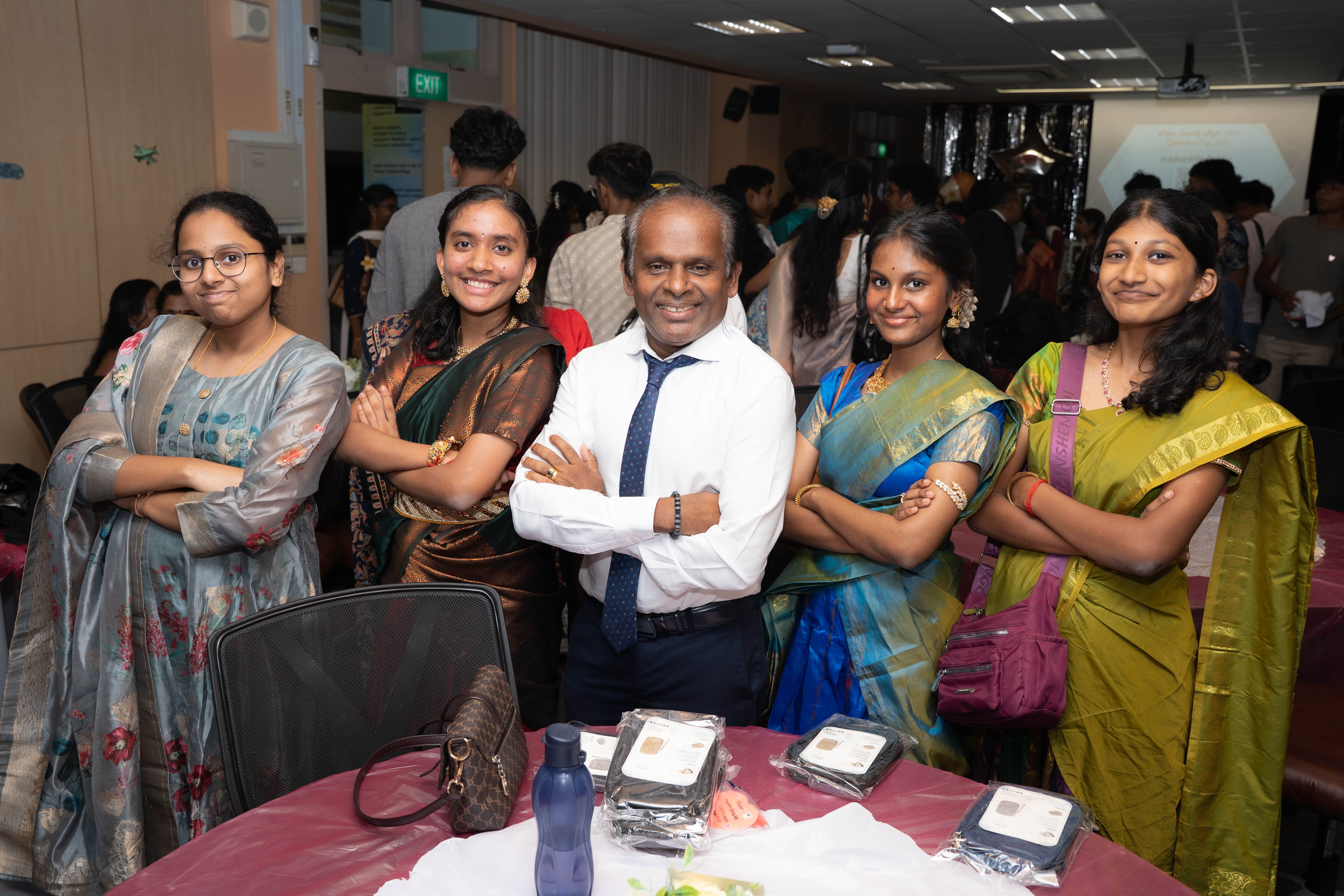 Five people smiling with arms crossed at an indoor event; four women in saris, one man in shirt and tie.