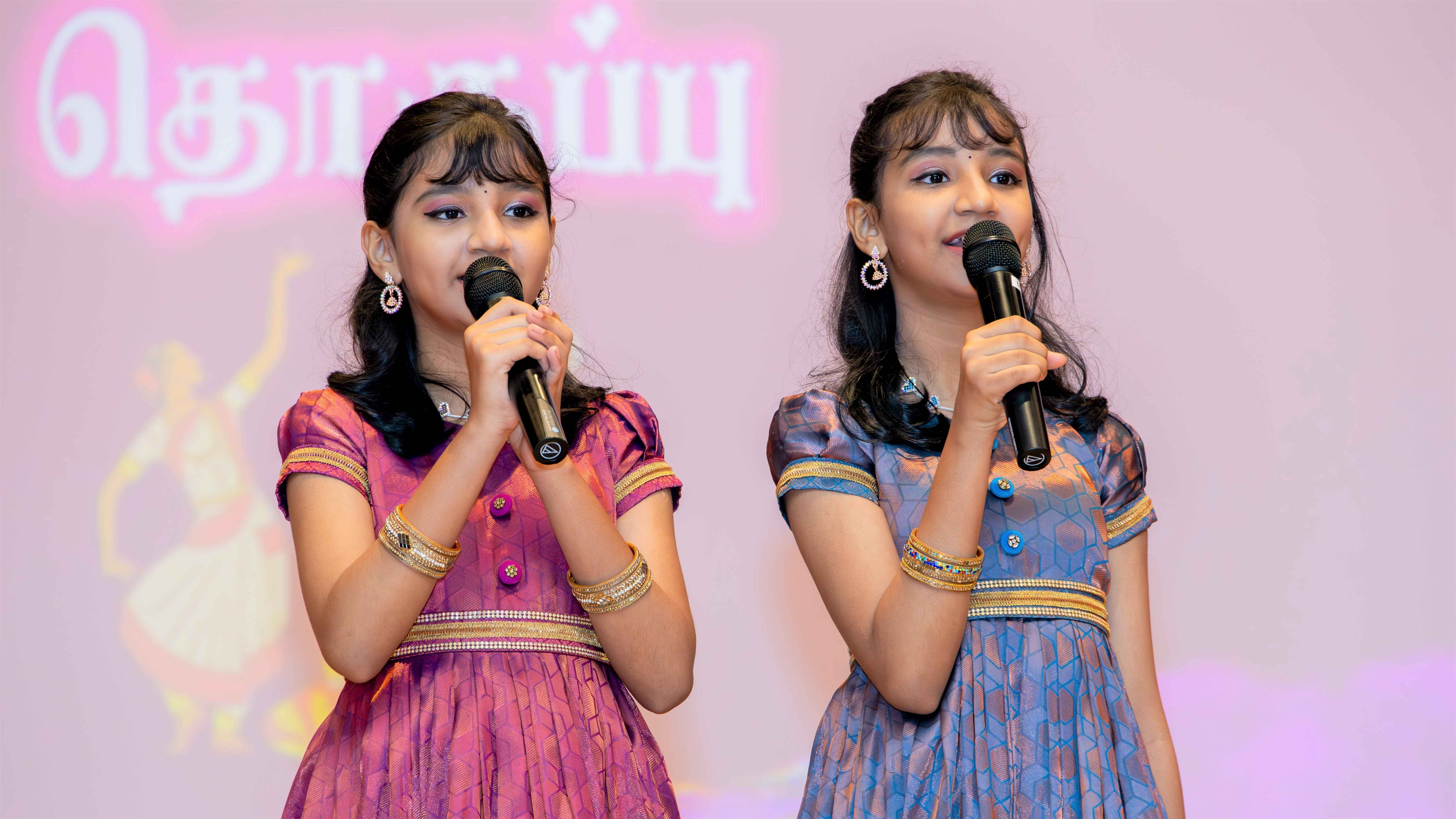 Two girls in colorful dresses hold microphones and sing on stage against a pink backdrop with Tamil text.