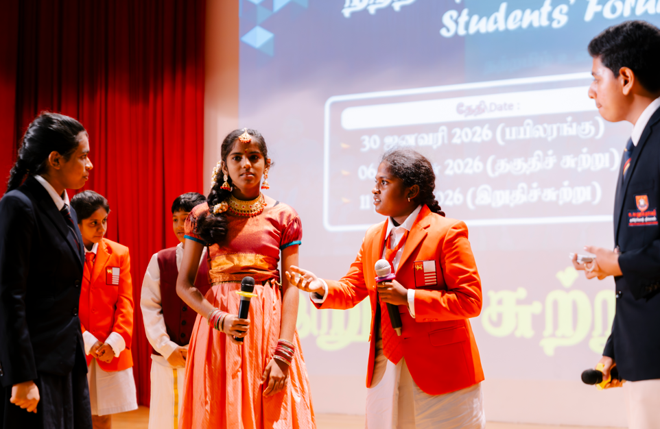 Students on stage; girl in orange dress holds mic while another in red blazer speaks into a mic.