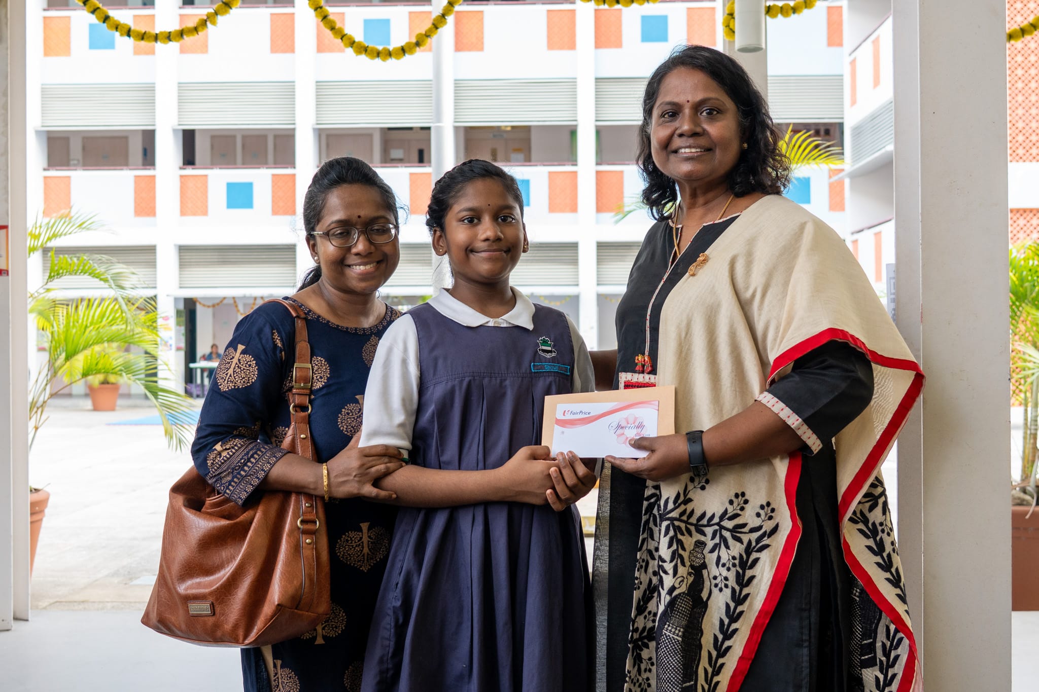 Woman in sari hands an envelope to a schoolgirl as another woman stands beside them in a courtyard.