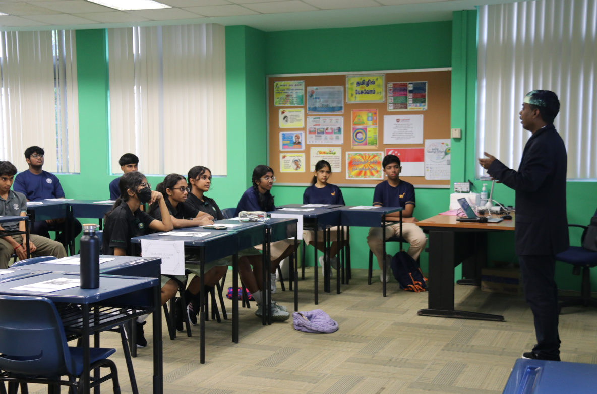 Teacher in suit speaks to seated students at desks in a green classroom with posters on a bulletin board.