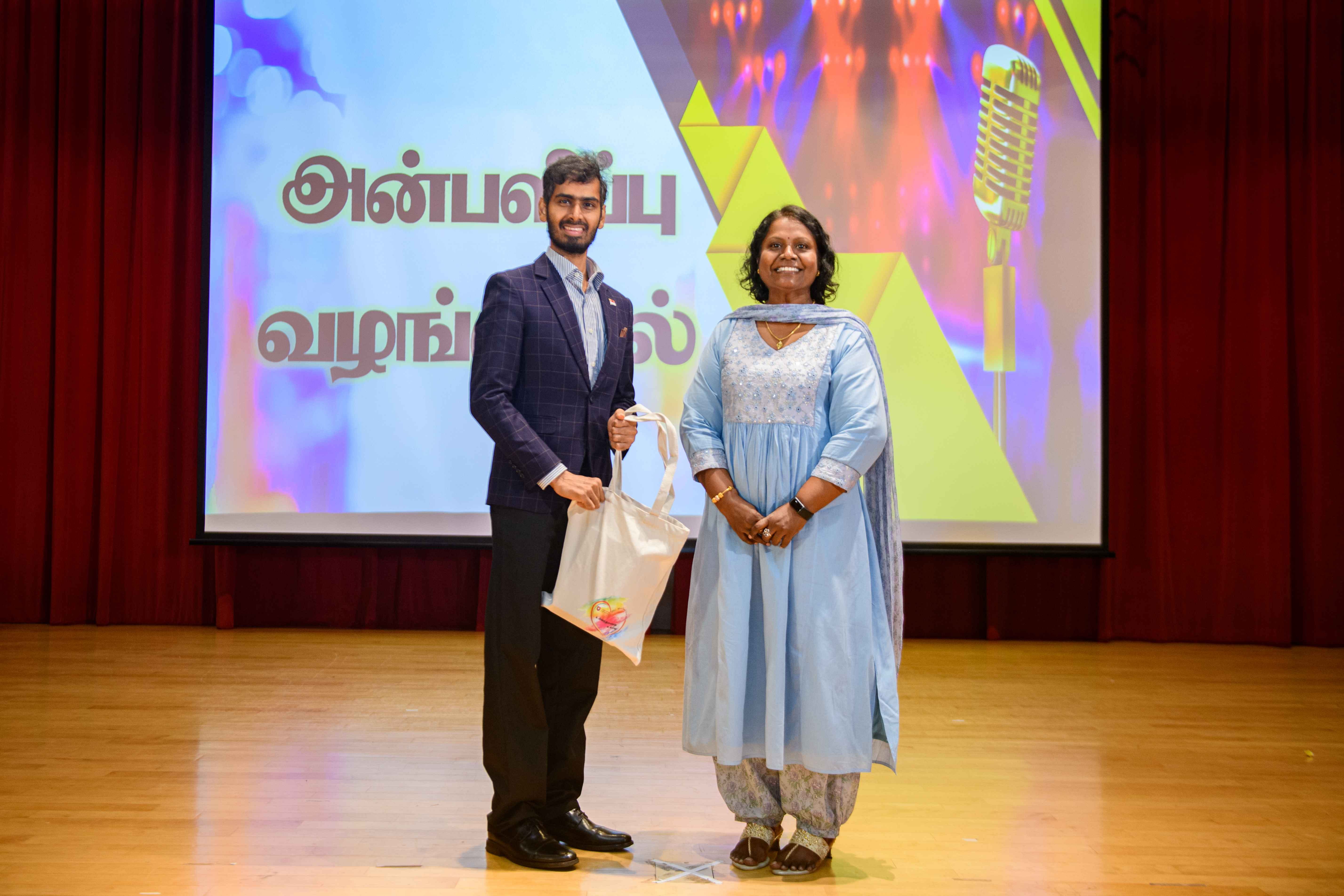 Man holding a white tote bag stands beside a woman in a blue dress on stage before a screen with Tamil text.