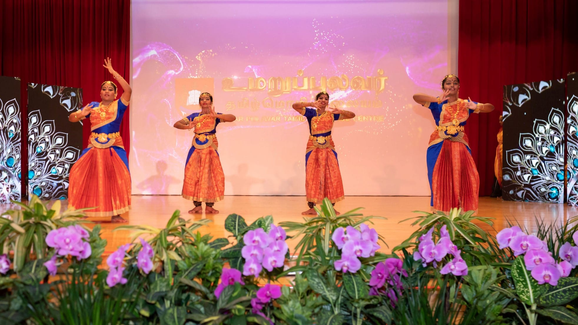 Four dancers in orange and blue costumes pose on a stage with red curtains and purple flowers in front.