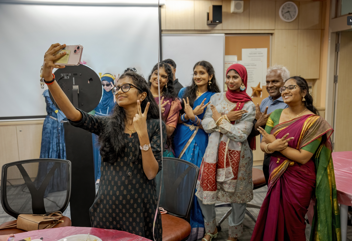 Woman takes a selfie flashing peace sign as a group in colorful outfits pose and smile in a room