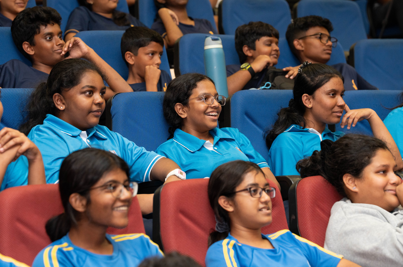 Students seated in an auditorium, many smiling and watching something off to the right.
