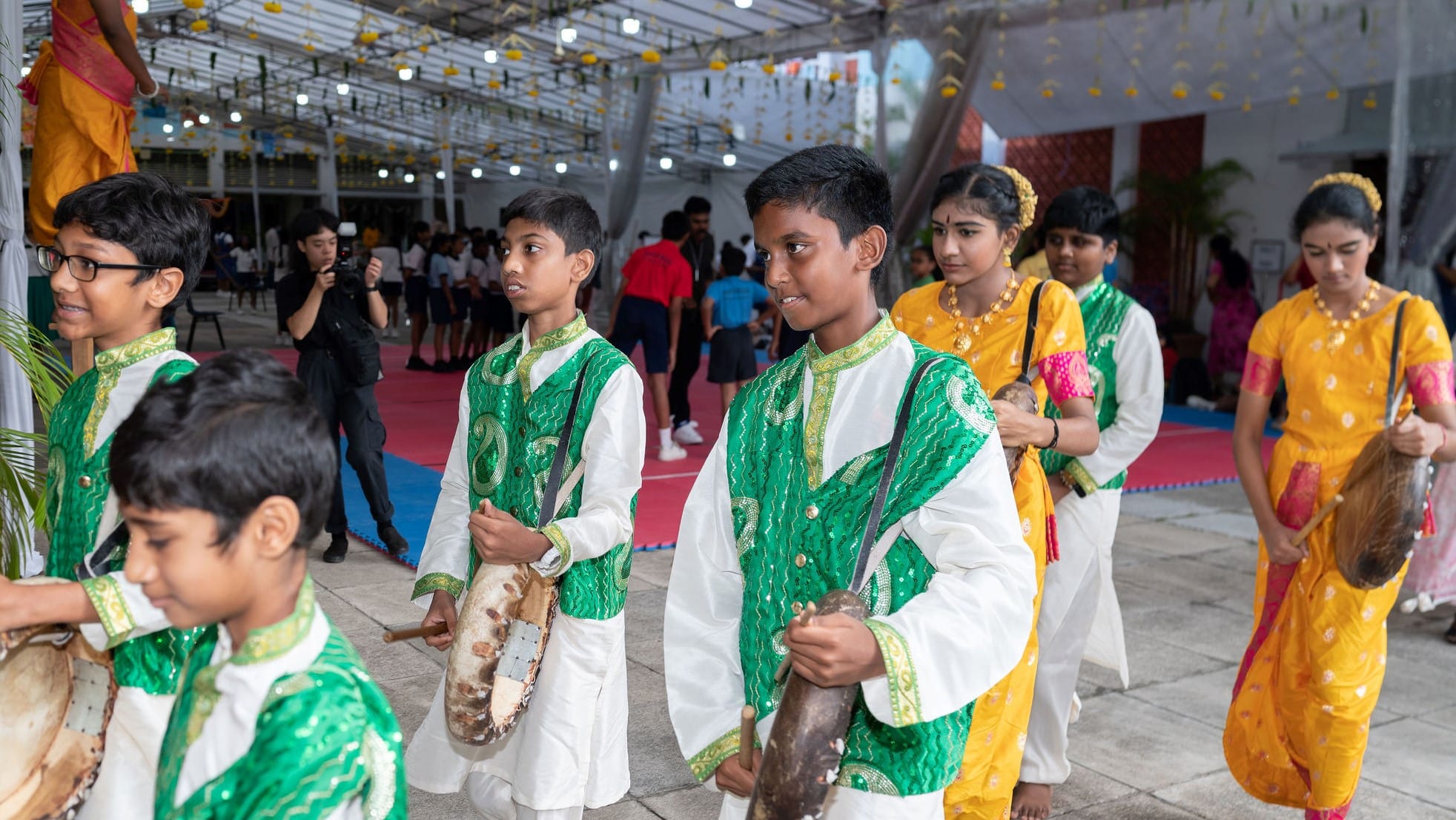 Students in colorful traditional outfits walk in a line carrying drums and cymbals under a decorated canopy.