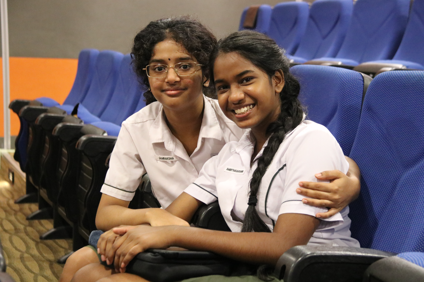 Two students in white uniforms sit in blue theater seats, smiling; one hugs the other from behind.