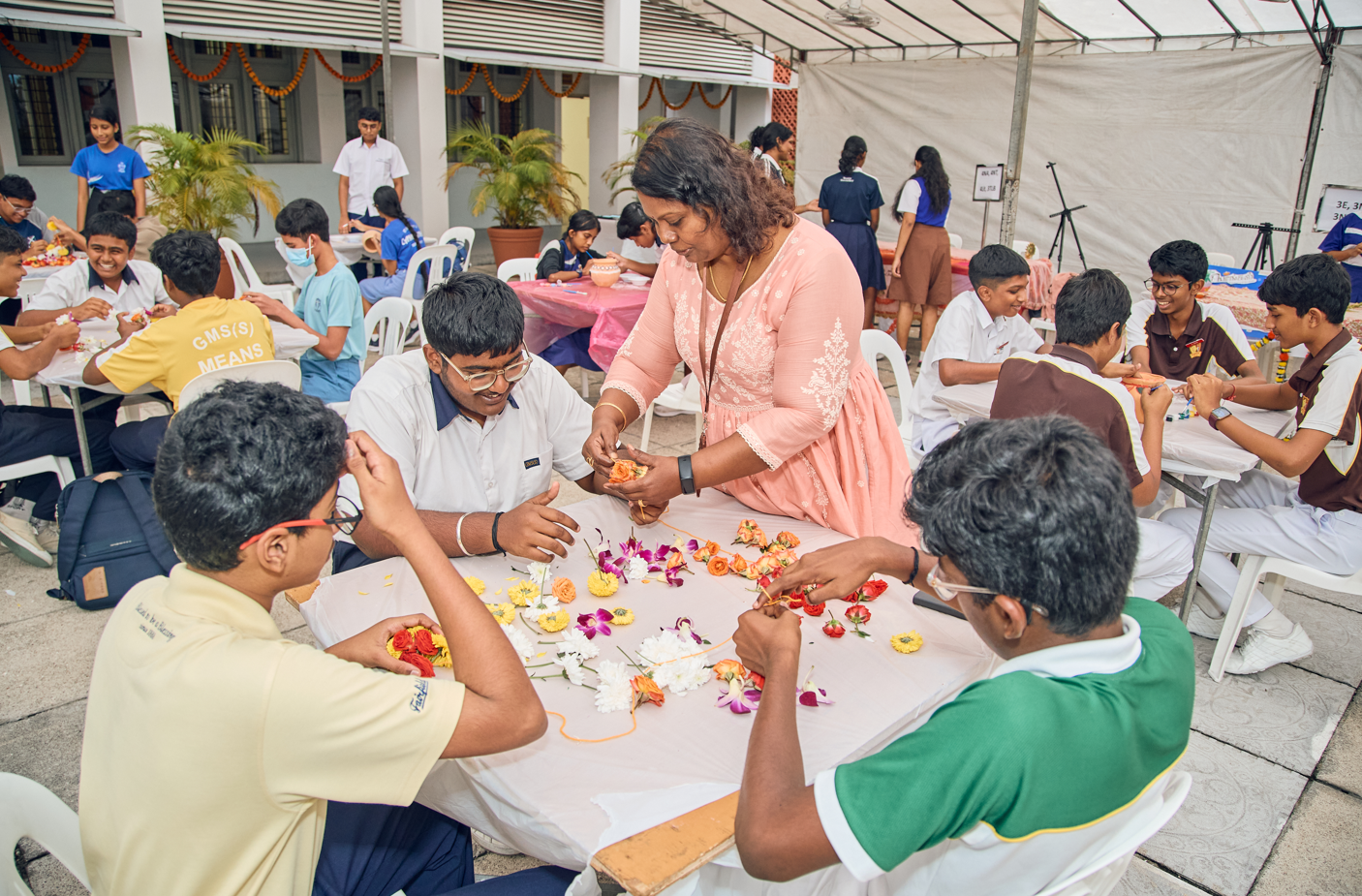 Teacher helps students arrange colorful flowers on a table in a busy school courtyard workshop.